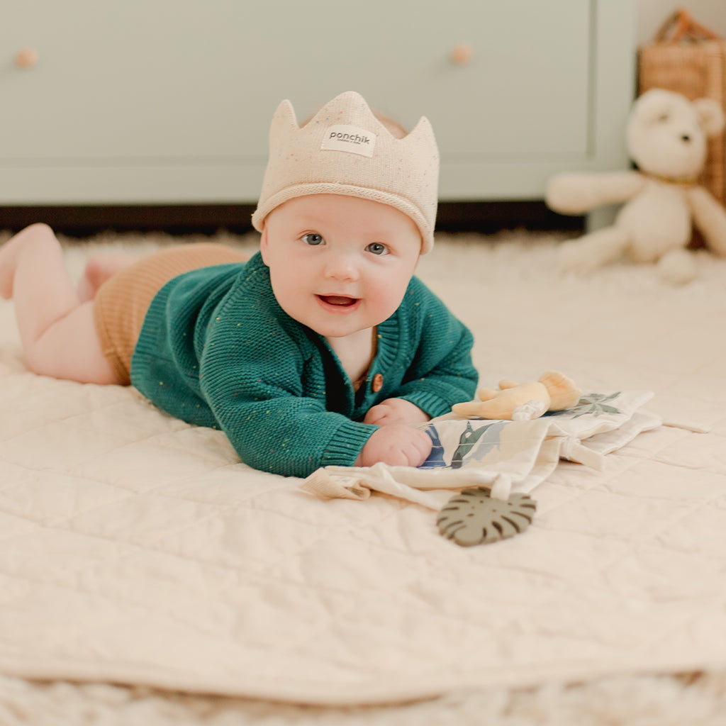 Baby lying on a play mat wearing a green sweater and beige crown, surrounded by toys.